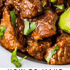 Close-up of tender beef chunks in a rich brown sauce, garnished with fresh cilantro and served on a white plate with sliced cucumber. Text overlay reads, “Tejano Carne Guisada.”.