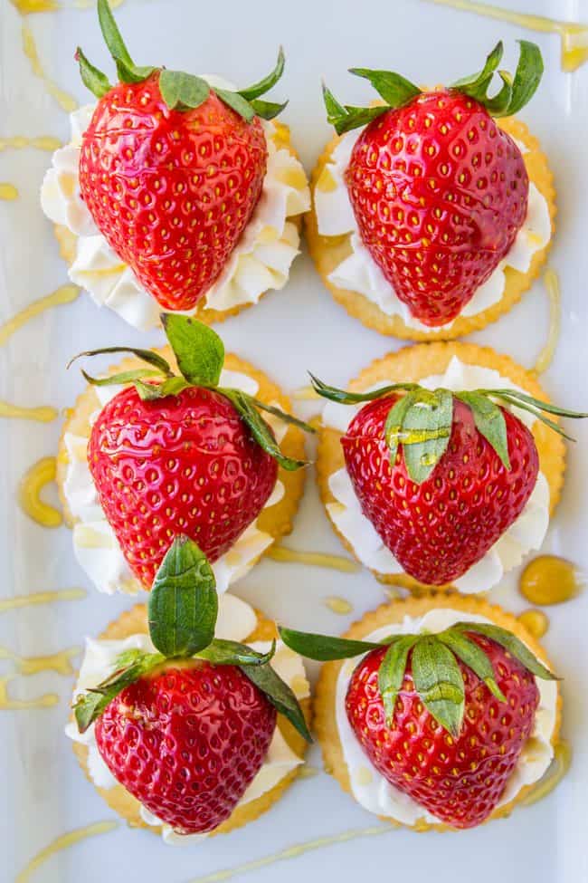 Six round crackers topped with swirls of whipped cream and large fresh strawberries, arranged neatly on a white plate—this delightful Strawberry and Cream Cheese Appetizer is finished with a tempting drizzle of honey in the background.