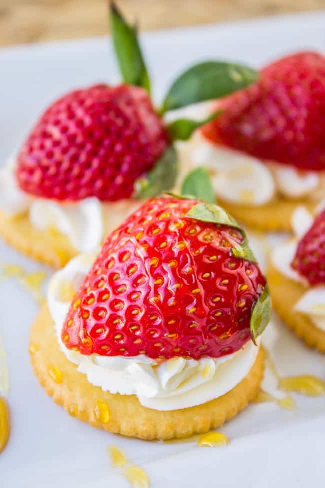 A close-up of a Strawberry and Cream Cheese Appetizer features a round cracker topped with a swirl of cream cheese, a fresh strawberry half, and a drizzle of honey. Other similar snacks are visible in the background.