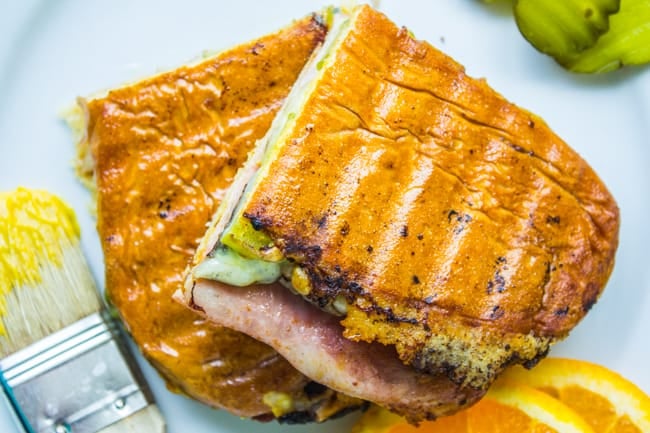 a Cuban sandwich cut in half with orange slices, pickles, and mustard nearby.