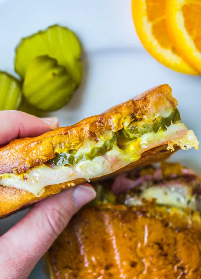 a hand holding a Cuban sandwich with pickles, cheese, pork, ham, and mustard.