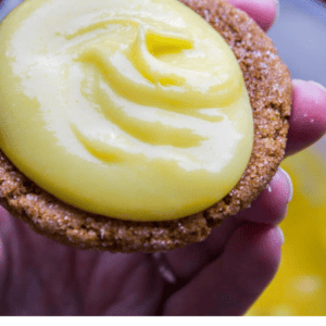 A hand holds a cookie topped with a generous swirl of creamy yellow lemon curd. Text below reads: The Very Best Lemon Curd, Quick & Easy, The Food Charlatan, featuring fresh lemon flavor.