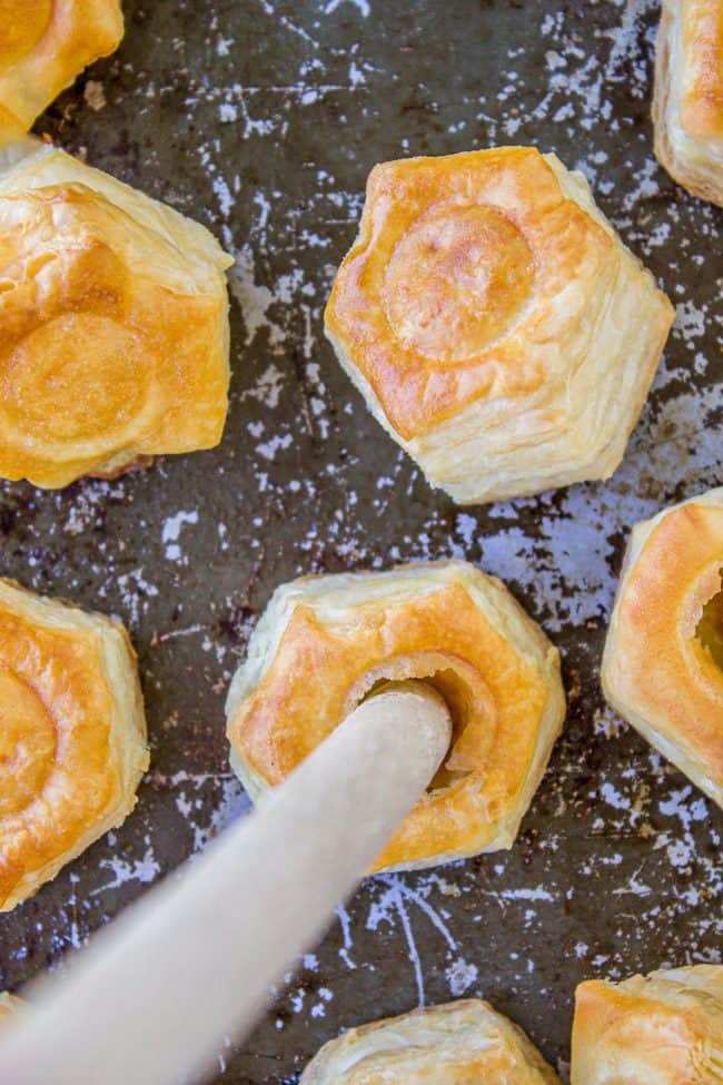 Hexagon-shaped pieces of golden puff pastry with hollow centers are arranged on a dark baking sheet. A wooden handle is inserted into the center of one pastry.