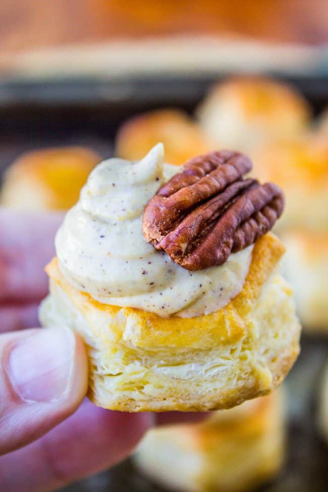 A hand holds a flaky pastry topped with creamy pumpkin spiced filling and a whole pecan, with more pastries blurred in the background.