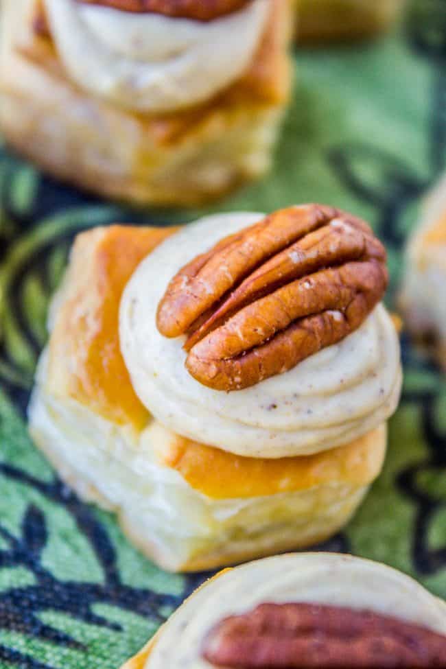 A close-up of a puff pastry bite topped with creamy pumpkin filling and a whole pecan, placed on a green patterned fabric.