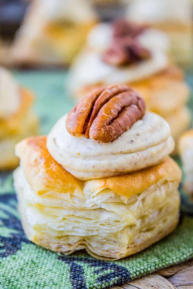 A close-up of a puff pastry bite topped with creamy pumpkin frosting and a pecan half, set on a green patterned cloth with more pastries blurred in the background.