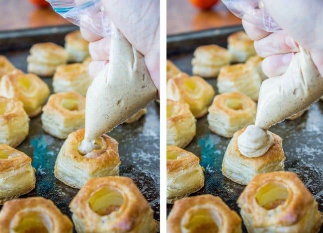 Close-up of a hand piping creamy filling into small, round, hollow puff pastry shells on a baking tray.