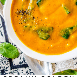 A bowl of creamy golden cauliflower soup with spinach leaves, topped with ground black pepper, rests on a patterned surface beside a silver spoon and fresh spinach leaves for a vibrant, comforting meal.
