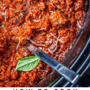 Close-up of rich, chunky spaghetti meat sauce simmering in a slow cooker, sprinkled with grated cheese and garnished with fresh basil leaves, with a serving spoon resting inside.