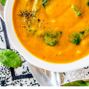 A bowl of creamy orange cauliflower soup with spinach leaves and black pepper on top, placed on a patterned cloth with fresh spinach nearby. Text reads: Easy 30 Minute Cauliflower Soup - The Food Charlatan.