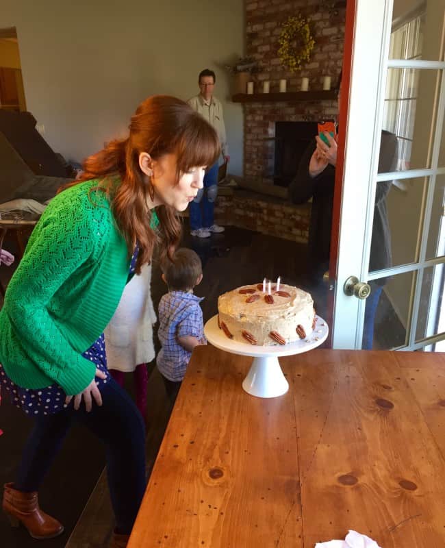 a redhead in a green sweater blowing out the candles on a carrot cake with pecans and cream cheese frosting. 