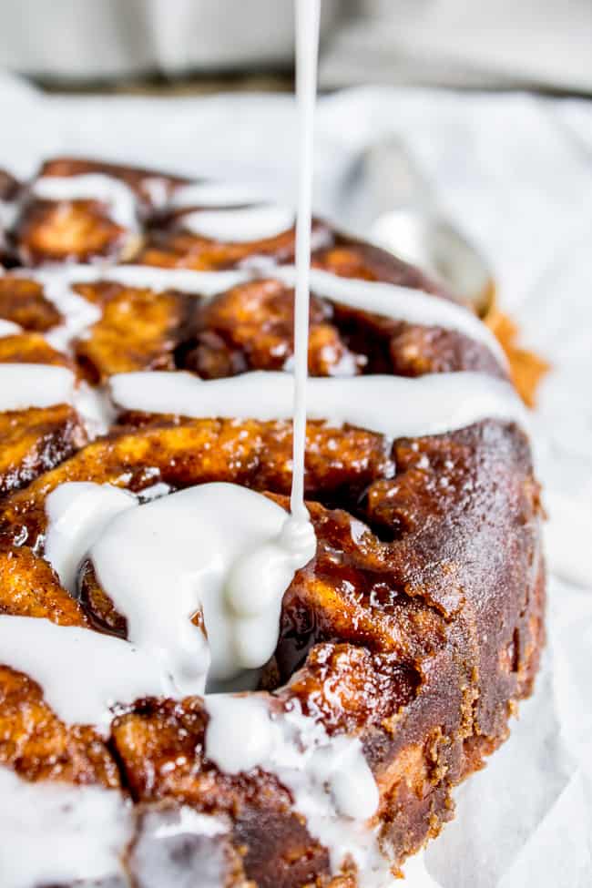 a stream of icing being drizzled onto a round cake pan of cinnamon rolls turned out onto parchment paper.