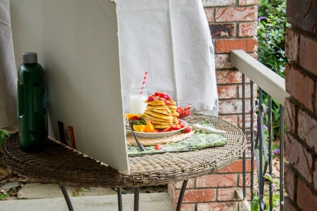 Pancake photo shoot setup showing a large, leaning stack of pancakes with fruit and syrup and a glass of milk with a striped straw.