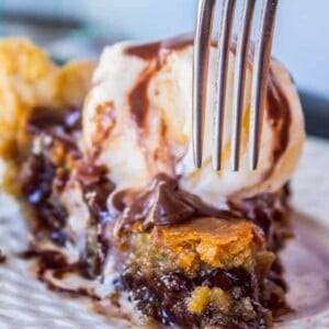 A fork cuts into a slice of gooey cookie pie topped with vanilla ice cream and drizzled with chocolate syrup, served on a white plate.