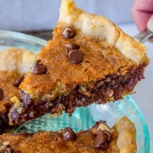 A slice of gooey cookie pie with a golden, flaky crust is being lifted from a glass pie dish. The pie is topped with melted chocolate chips.