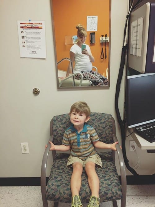 a little boy sitting in a chair at the doctor's office and his pregnant mother's reflection in the mirror.