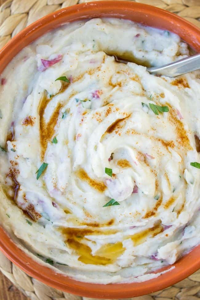 A bowl of creamy mashed potatoes garnished with chopped herbs, a sprinkle of paprika, and drizzled with melted butter, served alongside roasted potatoes and onions, with a spoon resting inside the bowl.