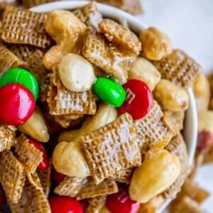 A close-up of a bowl filled with caramel-coated Christmas Chex Mix, cashews, almonds, and red and green candy-coated chocolates, with some spilling onto a white surface.