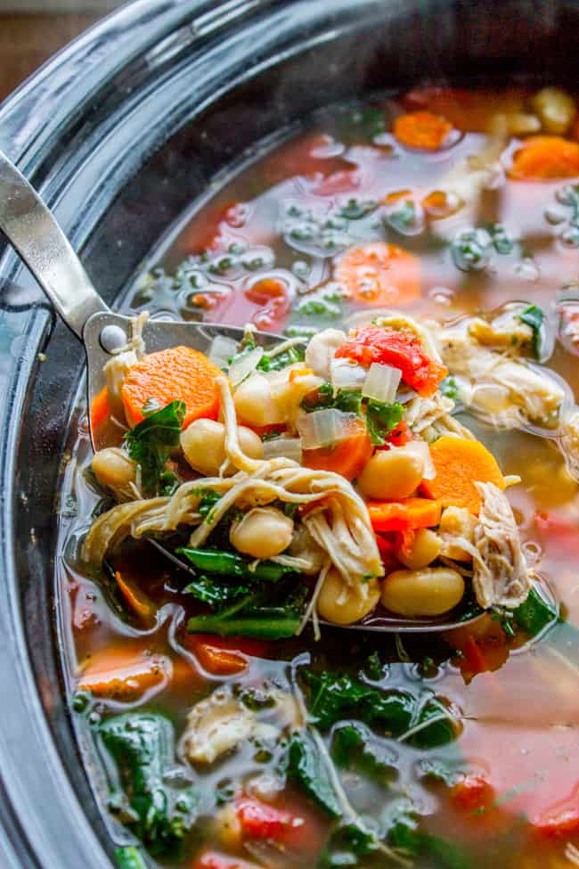 a spoon lifting chicken, kale, and white bean soup from a slow cooker.