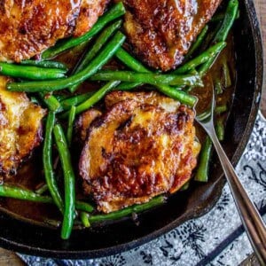 A cast iron skillet filled with chicken and green beans in a savory glaze sits on a wooden table. Text at the bottom reads, Asian Chicken with Green Beans.