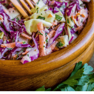 A wooden bowl filled with creamy coleslaw made of shredded cabbage, carrots, and parsley, with a gold fork in the bowl. Text below reads: Seriously the best coleslaw you’ll ever have – The Food Charlatan. Perfect for summer gatherings.