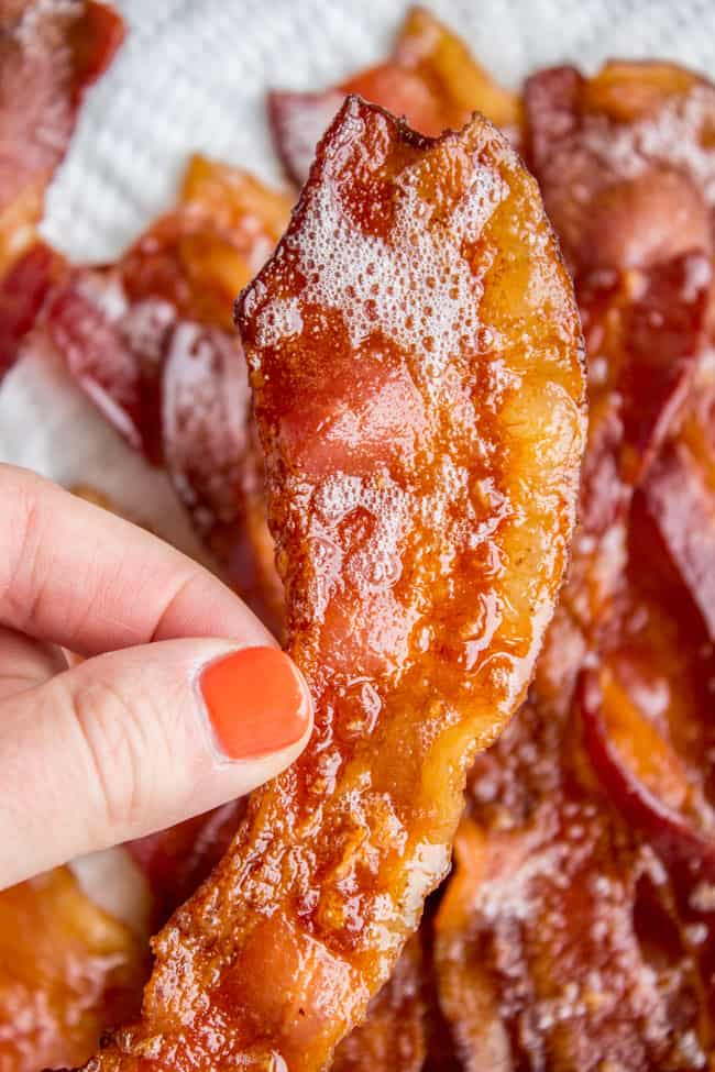 a hand holding a strip of crispy bacon above other strips on a sheet pan.