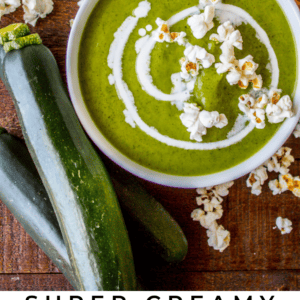 A bowl of creamy green zucchini soup topped with a swirl of cream and popcorn sits beside fresh zucchinis and scattered popcorn, with the text “Super Creamy Zucchini Soup” at the bottom, highlighting its deliciously smooth texture.