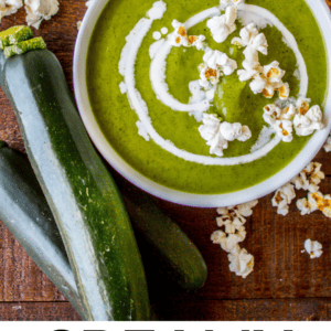 A bowl of creamy green zucchini soup topped with a swirl of cream and popcorn sits on a wooden table beside fresh zucchinis, a green towel, and extra popcorn. Text reads Creamy Zucchini Soup for a cozy mealtime delight.