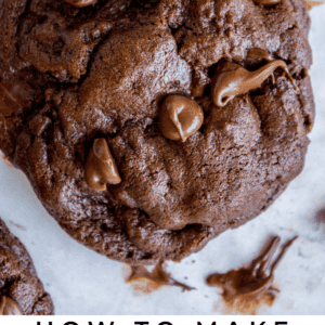 Close-up of a thick double chocolate chip cookie packed with visible chocolate chips, surrounded by more cookies and melted chocolate on a light surface. Text reads: Double Chocolate Chip Cookies. The Food Charlatan.