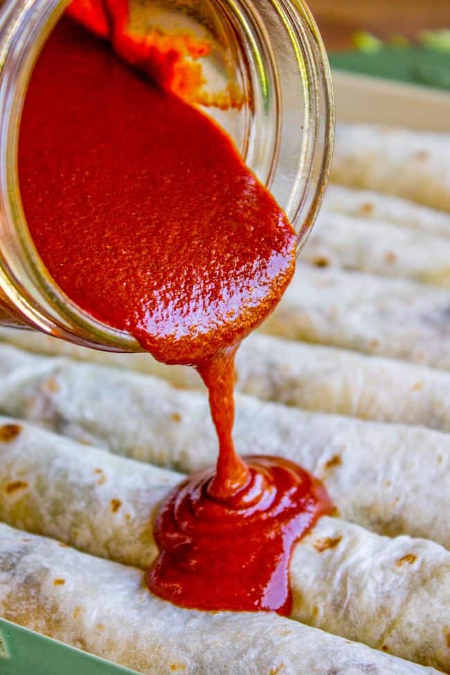 pouring red enchilada sauce onto a pan of enchiladas.