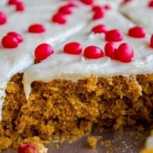 A close-up of moist, sliced pumpkin bars topped with white frosting and scattered red candies, with one piece partially removed from the pan.