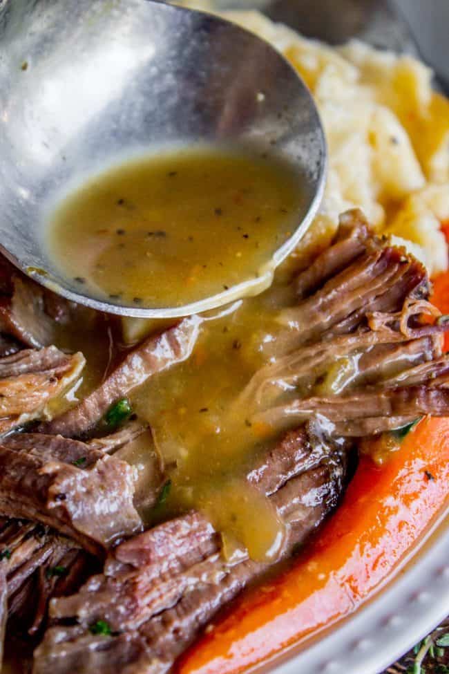 pouring gravy from a ladle over crockpot pot roast, carrots, and mashed potatoes.