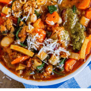 A bowl of hearty slow cooker minestrone with Italian sausage, pasta, beans, carrots, spinach, and zucchini, finished with a dollop of pesto and shredded cheese, served with a spoon on the side.