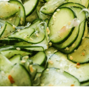 Thinly sliced cucumbers tossed with sesame seeds and a savory dressing, served in a bowl. Text overlay reads: “Light & Refreshing Asian Cucumber Salad—The Food Charlatan.”.