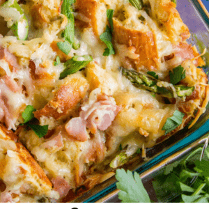Close-up of savory bread pudding in a glass dish, featuring chopped ham, asparagus, melted cheese, and fresh parsley. The text below reads: Savory Bread Pudding with Ham & Asparagus. The Food Charlatan.