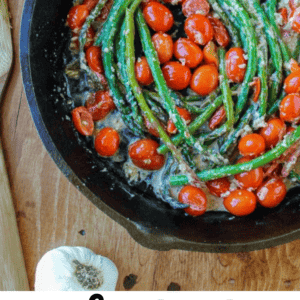 A cast-iron skillet with sautéed asparagus and cherry tomatoes in a creamy sauce sits on a wooden surface near garlic, salt, and a wooden spoon. Vibrant cherry tomatoes add color and flavor to this delicious sautéed asparagus dish. Text: Sautéed Asparagus with Cherry Tomatoes.