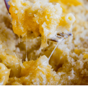 A close-up of creamy, cheesy macaroni and cheese being scooped out with a fork, topped with a golden breadcrumb crust. Text below reads: Literally the cheesiest mac and cheese I’ve ever had! The Food Charlatan.