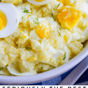 A close-up of classic potato salad topped with sliced hard-boiled eggs and fresh dill, served in a white bowl. Vibrant text at the bottom reads: Seriously the Best Classic Potato Salad. The Food Charlatan.