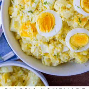 A bowl of classic homemade potato salad, topped with sliced hard-boiled eggs and fresh dill, sits on a table beside a napkin and basket. Text below: Classic Homemade Potato Salad with no crunchies! The Food Charlatan.