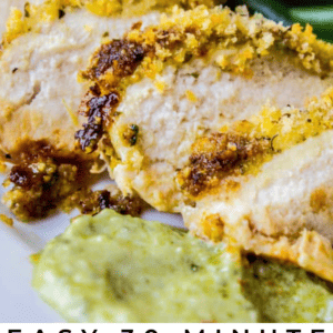 Close-up of sliced baked chicken breast topped with golden breadcrumbs and vibrant pesto, served with green beans and extra pesto sauce. Text overlay reads: Easy 30 Minute Baked Pesto Chicken. The Food Charlatan.