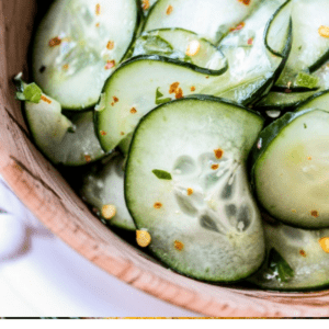 Close-up of a wooden bowl filled with cucumber slices garnished with fresh cilantro, lime, and red pepper flakes. Text below reads, Quick & Easy Cilantro-Lime Cucumber Salad. The Food Charlatan.