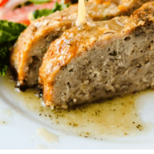 Close-up of two thick slices of meatloaf topped with garlic butter sauce being poured from a spoon, served with a fresh side salad. Text overlay: 1770 House Meatloaf with Garlic Butter Sauce - The Food Charlatan. Classic comfort food at its finest.