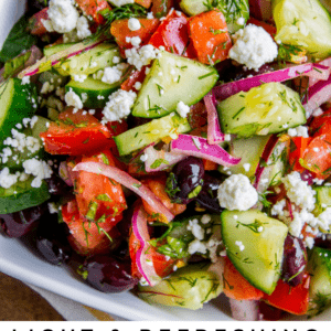 A close-up of a colorful Greek salad with tomatoes, cucumbers, red onions, olives, feta cheese, and fresh herbs in a white bowl. Text on image: Light & Refreshing Greek Salad – The Food Charlatan.