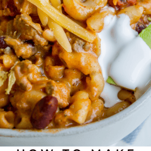 Close-up of a bowl of chili mac and cheese topped with shredded cheese, sour cream, and green onions, with a spoon. Text at the bottom reads âChili Mac & Cheese. The Food Charlatan.â.