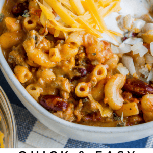 A bowl of chili mac and cheese topped with shredded cheddar, chopped onions, and a dollop of sour cream. Text below reads: Quick & Easy Chili Mac & Cheese - The Food Charlatan, featuring hearty comfort food flavor.