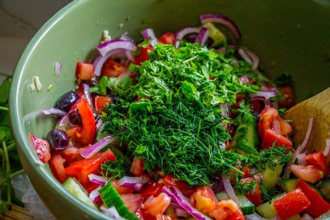 mixing fresh dill and mint with other ingredients to make Greek cucumber tomato salad with feta cheese in a  green mixing bowl. 