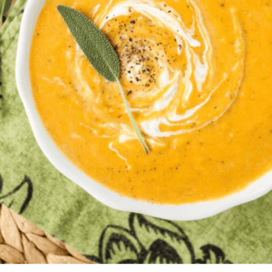 A bowl of creamy butternut squash soup garnished with a swirl of cream, black pepper, and a sage leaf sits on a green patterned cloth. Text reads: Classic Butternut Squash Soup - The Food Charlatan.