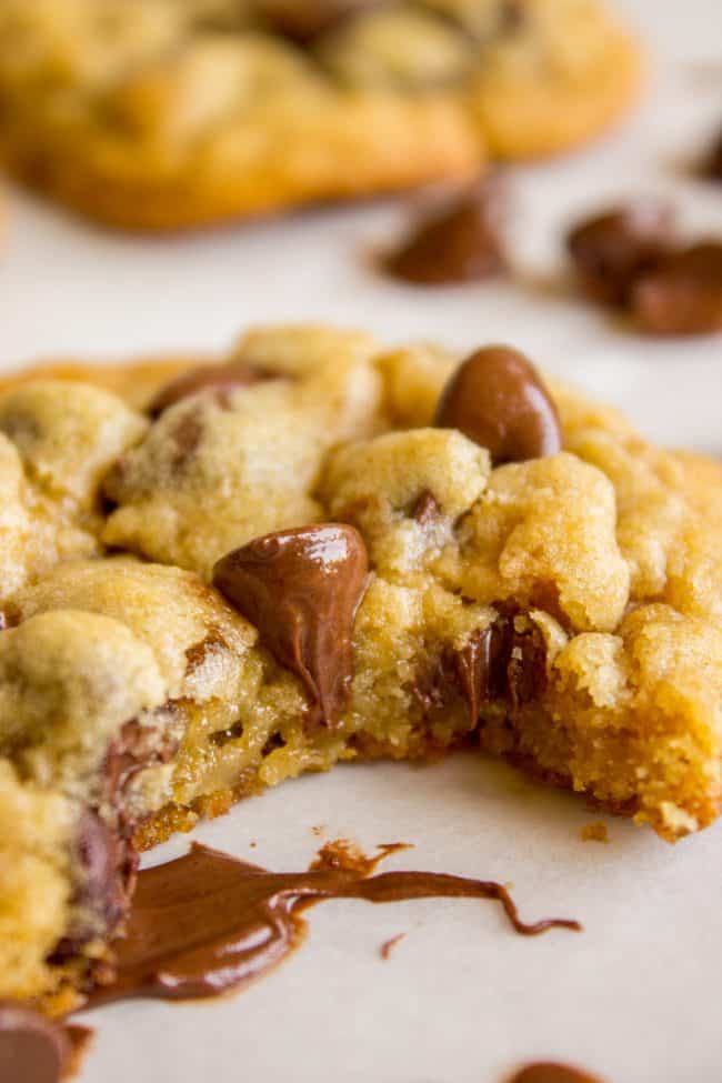 a close up shot of a chocolate chip cookie with a bite taken out of it and chips melting onto parchment paper.