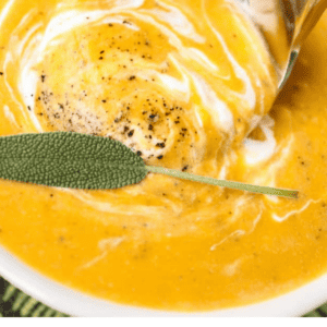 A bowl of creamy roasted butternut squash soup topped with a swirl of cream, black pepper, and a sage leaf, with a spoon scooping up the rich, velvety soup.