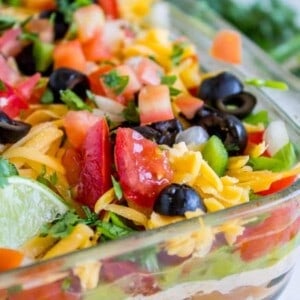 A close-up of a glass dish filled with 7 layer dip, topped with shredded cheese, diced tomatoes, sliced black olives, chopped green onions, and cilantro. A lime wedge is visible on the side.
